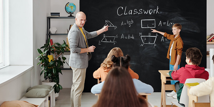 Math teacher and student pointing at shapes on a blackboard in front of a class of school children.