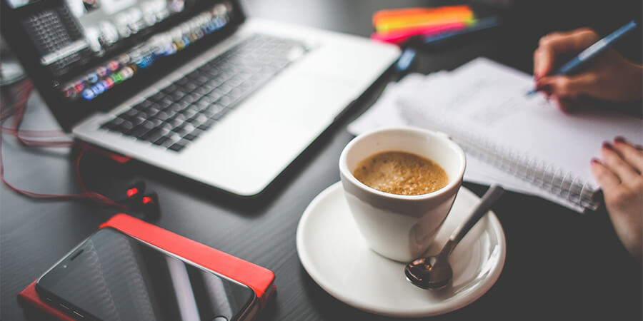 Desk with woman's hand writing in notebook and a smart phone, laptop and espresso.