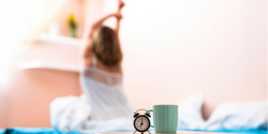 Alarm clock and coffee mug with woman stretching in the background.